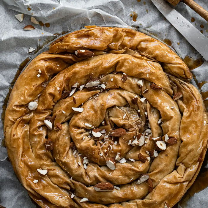 Auf einem Backpapier liegt ein Kaffee Baklava, daneben finden sich zerhackte Mandeln, ein Messer und ein Pinsel 