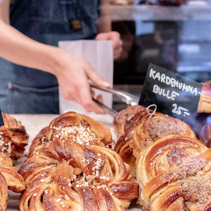 Im Vordergrund liegen Kanelbulle, im Hintergrund greift eine Frau mit einer Zange eine Kanelbulle an, um sie in ein Papiersäckchen zu geben