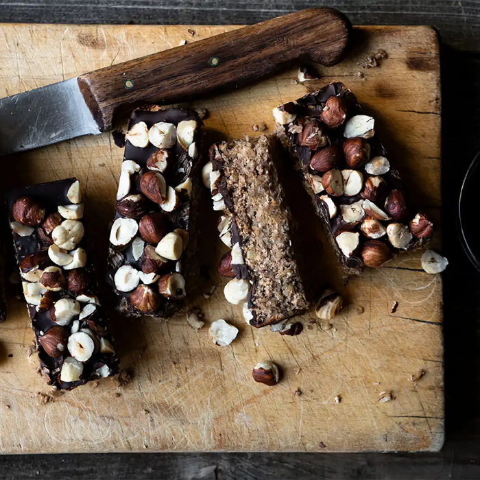 In Stücke geschnittene Coffee-Oat-Bars auf einem Brett mit einem Messer, zerkleinerten Haselnüssen und einer Tasse Kaffee von oben 
