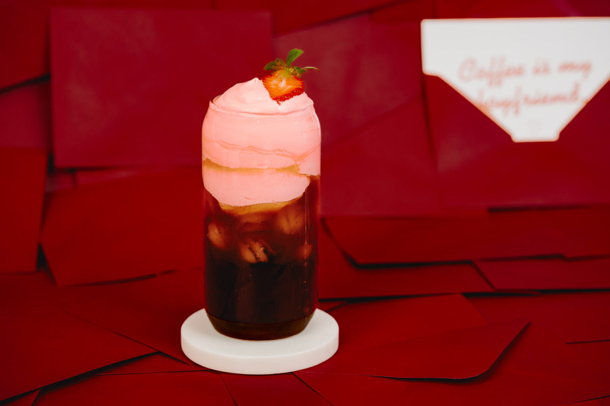 Der Cold Brew mit Strawberry Cold Foam ist in einem Glas von der Seite zu sehen. Das Glas steht auf einem hellen Untersetzter vor rotem Hintergrund.