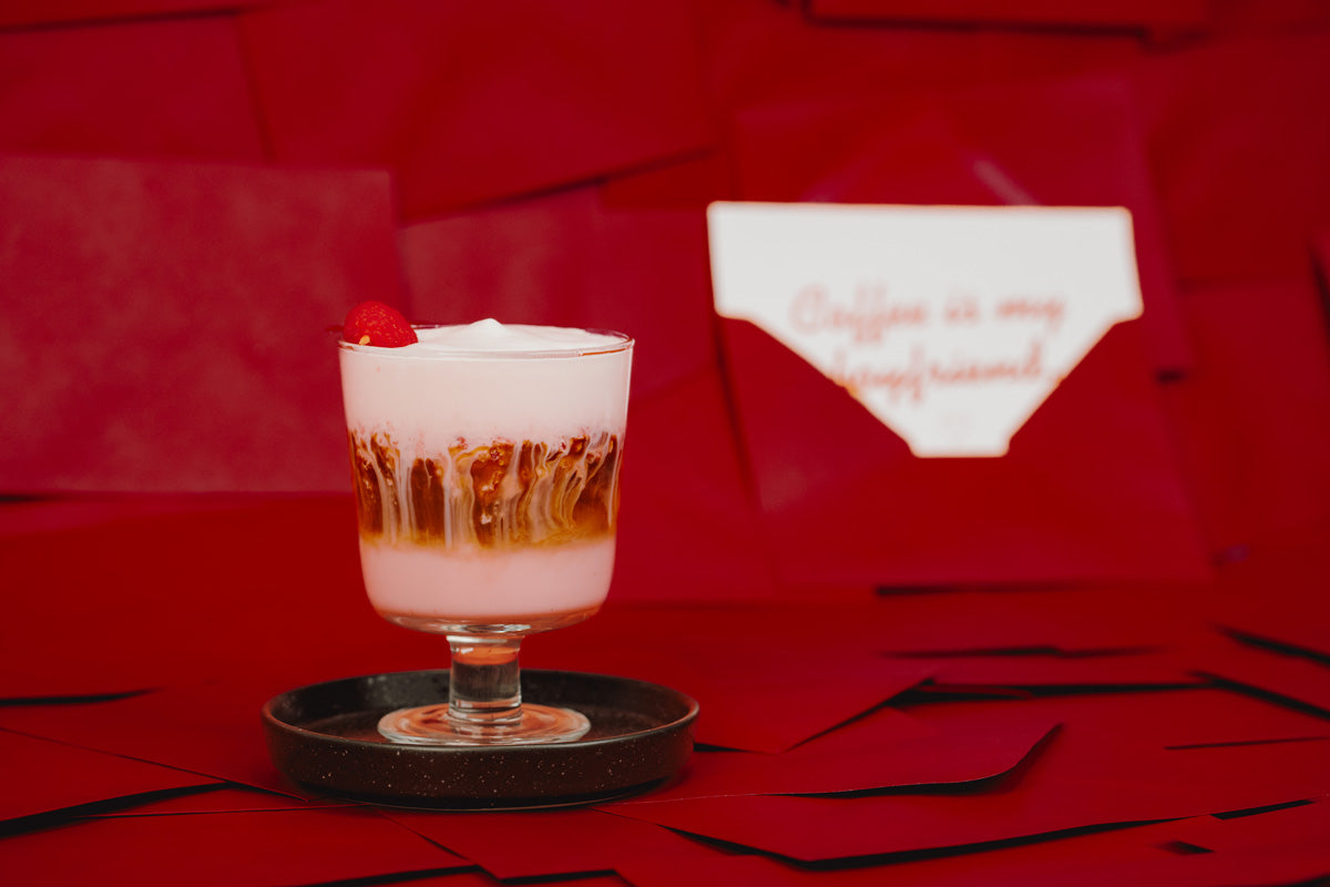 Der Raspberry Latte ist in einem Glas von der Seite zu sehen. Das Glas steht auf einem dunklen Untersetzter mit rotem Hintergrund.
