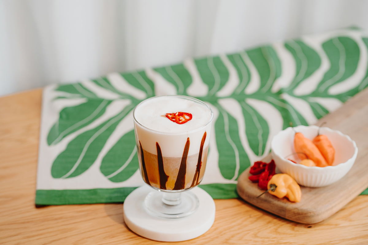 Ein Glas gefüllt mit Chili-Schoko-Kaffee steht am Tresen. Daneben liegen Chilischoten auf einem Holzbrett.