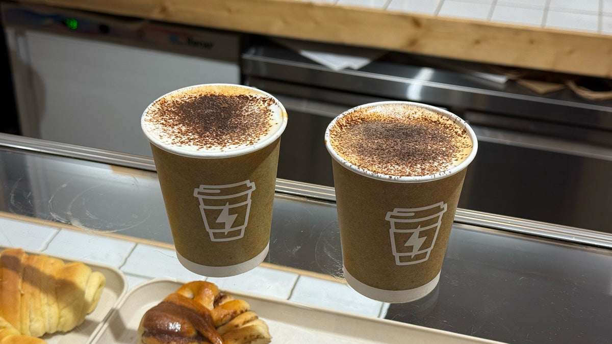 Zwei To-go-Cappuccinos, mit Kakaopulver bestäubt, stehen nebeneinander auf dem Tresen.