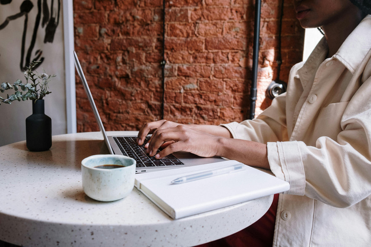 Eine Frau trinkt einen Kaffee an einem Tisch. Sie arbeitet an einem Laptop.