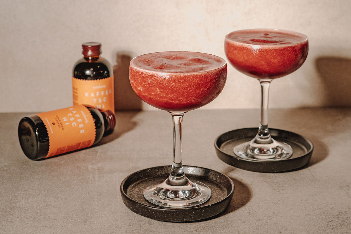 Zwei rote Frozen Cherry Espresso Martinis  in Coupé-Gläsern mit J. Hornig Kaffeetschi im Hintergrund.