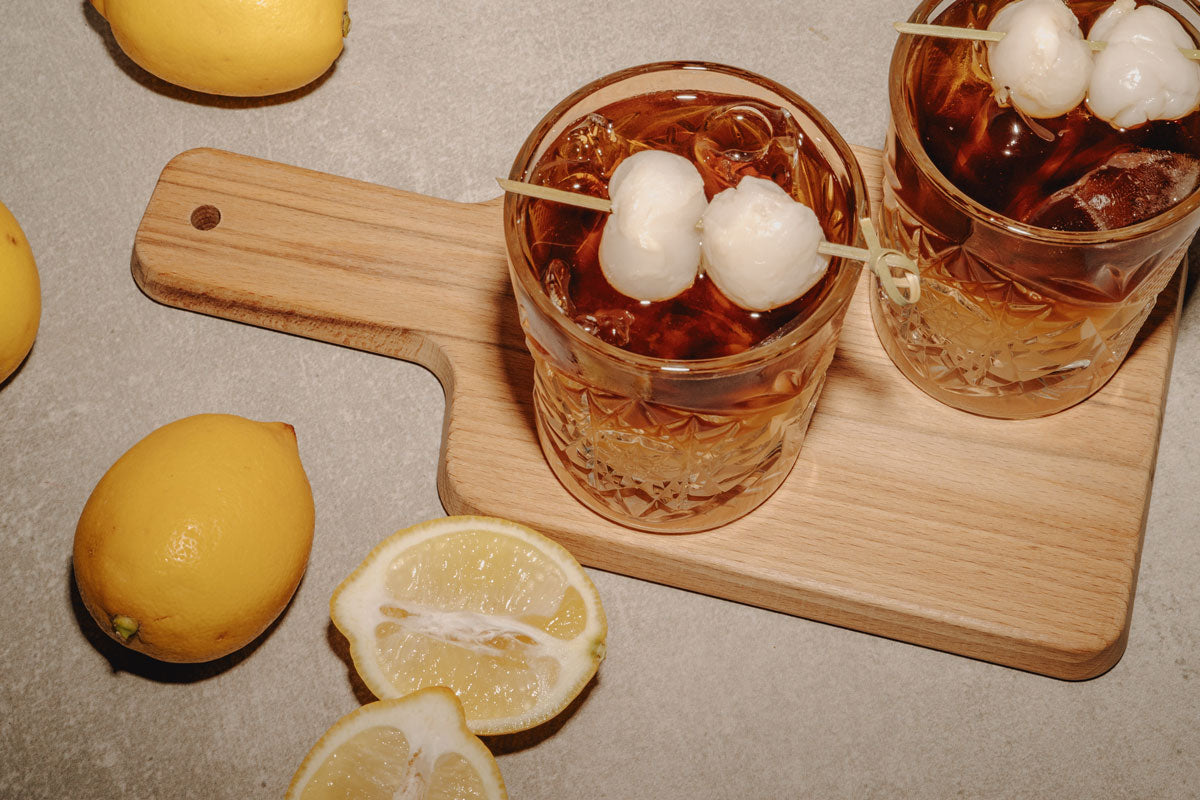 Zwei Lychee Cider Cold Brew Drinks mit Litschi-Garnitur und Zitronen auf einem Holzbrett.