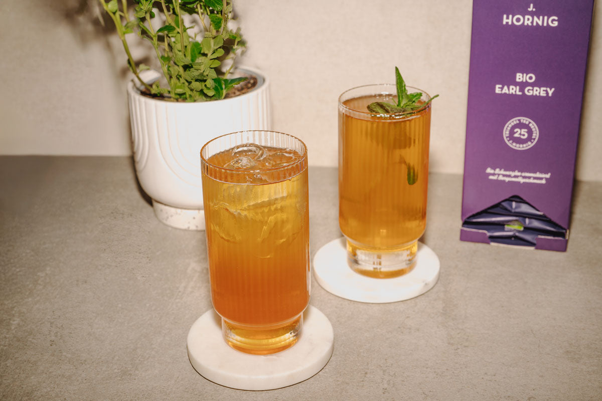 Zwei Gläser Earl Grey Gin Fizz mit Eiswürfeln und Minze neben einer Packung J. Hornig Bio Earl Grey Tee.