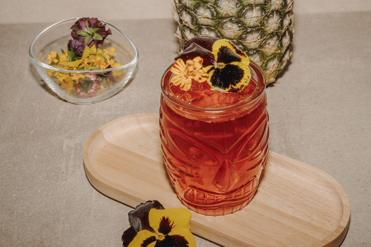 Hawaii Cocktail im Tiki-Glas mit essbaren Blüten und Ananas im Hintergrund.