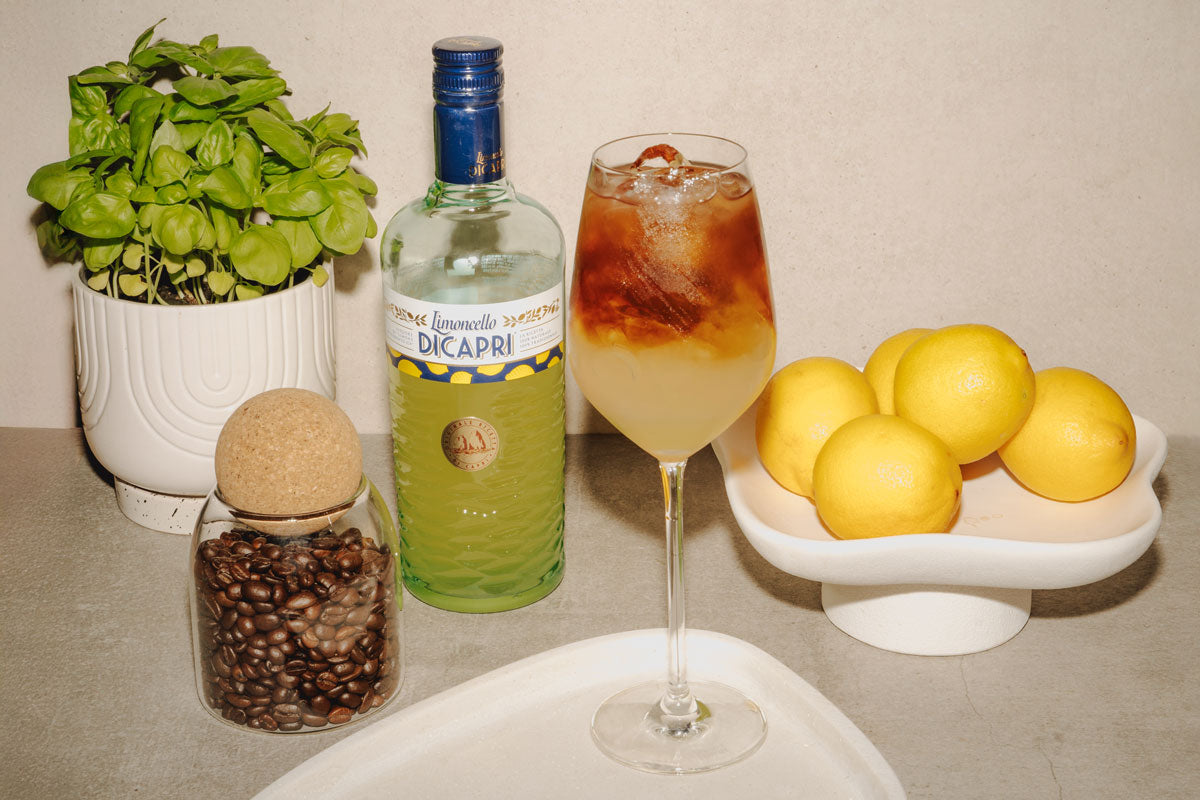 Limoncello-Kaffee-Cocktail mit J. Hornig Kaffeetschi im Weinglas, flankiert von Zitronen, Basilikum und Kaffeebohnen.