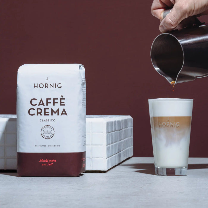 Eine Packung J. Hornig Caffe Crema Classico mit einem Latte Machiato Glas und einer Kanne auf weißem Untergrund mit braunem Hintergrund