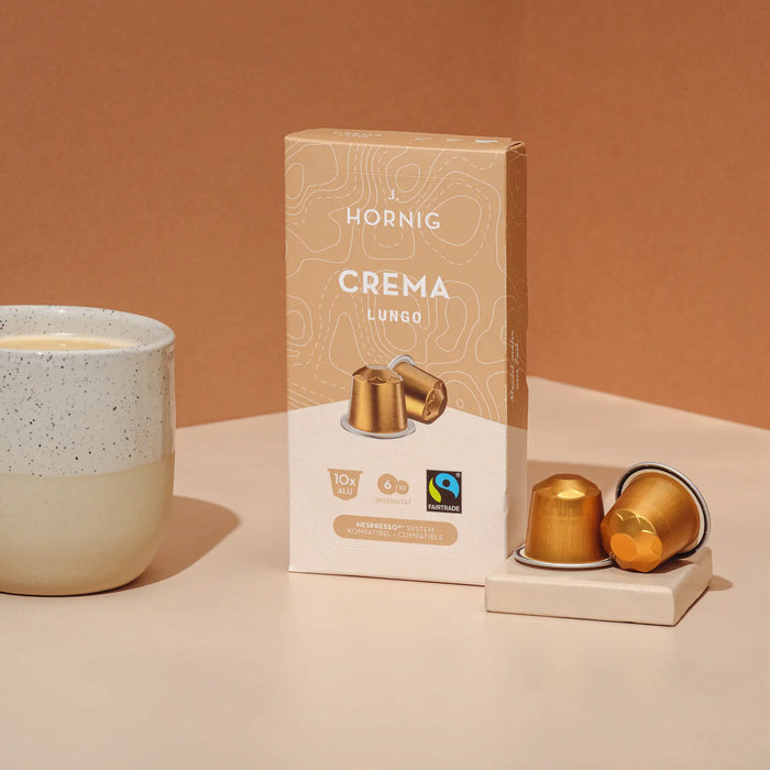 Eine Packung J. Hornig Crema Lungo Kapseln mit einer Kaffeetasse und zwei Alukapseln auf hellem Untergrund mit beigem Hintergrund.