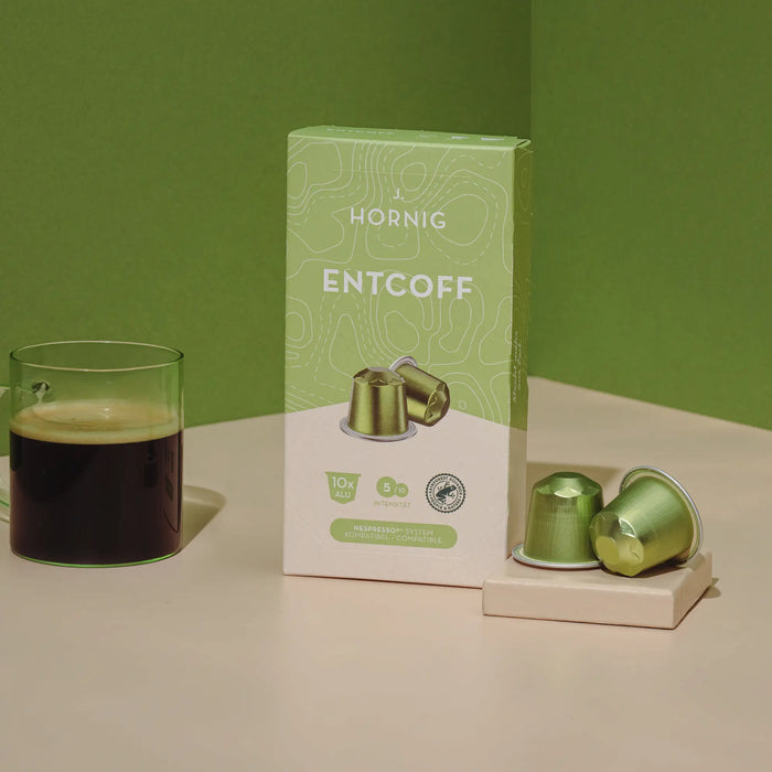 Eine Packung J. Hornig Entcoff Kapseln mit einer Kaffeetasse und zwei Alukapseln auf hellem Untergrund mit grünem Hintergrund.