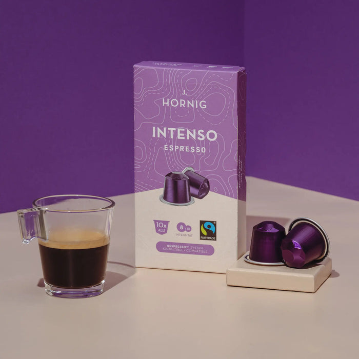 Eine Packung J. Hornig Intenso Espresso Kaffeekapseln mit zwei Alukapseln und einer Kaffeetasse auf hellem Untergrund mit einem grauen Hintergrund
