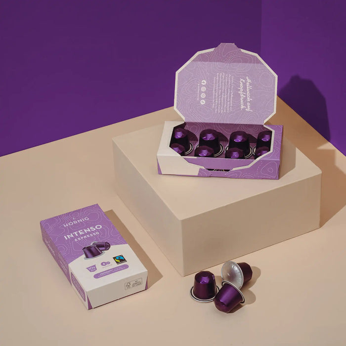 Eine offene Packung J. Hornig Intenso Espresso Kaffeekapseln und eine geschlossene Packung J. Hornig Intenso Espresso Kaffeekapseln auf einem hellen Untergrund mit einem violetten Hintergrund