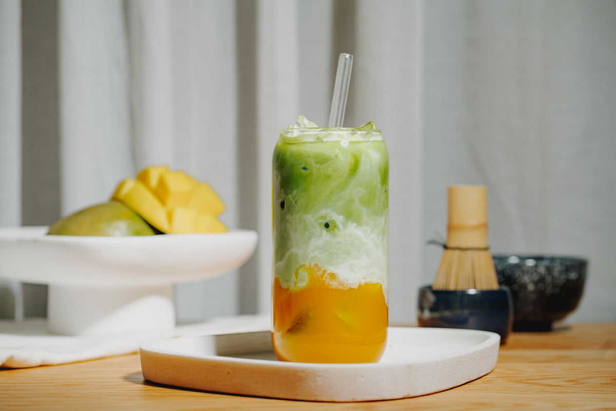 Ein Glas gefüllt mit Mango Matcha Iced Latte, im Hintergrund frische Mango und Matcha Set. 