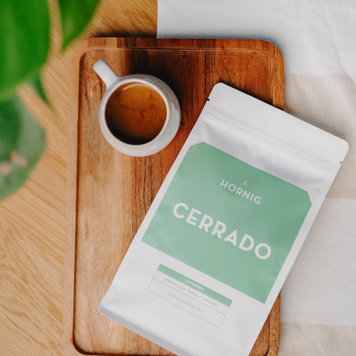 Eine Packung J. Hornig Spezialitätenkaffee Cerrado Espresso mit einer Espressotasse und einer Zimmerpflanze auf einem Holztisch