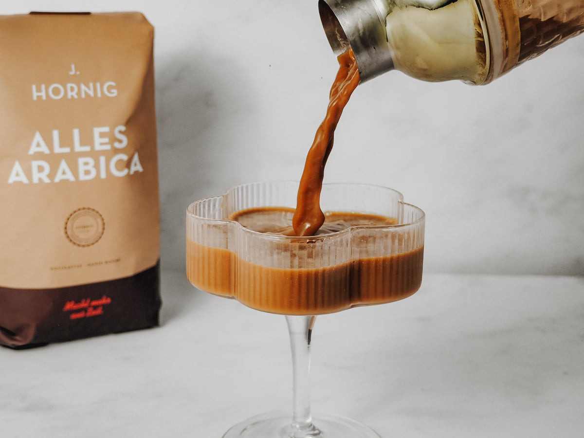 Tiramisu Espresso Martini wird in ein Martini Glas gegossen. Im Hintergrund steht eine Packung Alles Arabica von J. Hornig. 