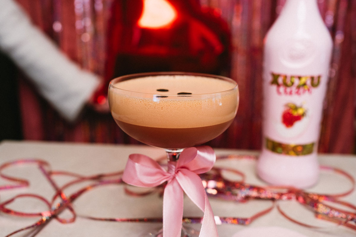 Pinker Espresso Martini steht auf einem Tisch, Hintergrund Frau, welche einen Herz-Ballon hält. 